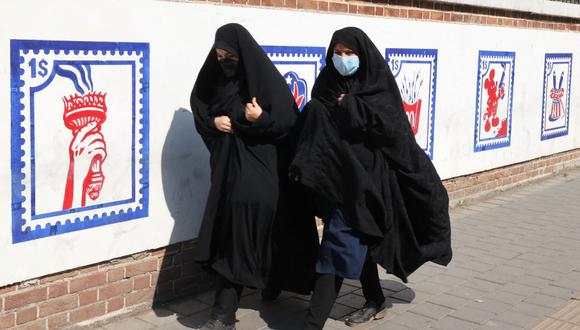 Mujeres con máscaras médicas pasan frente a un mural que representa diseños simulados de sellos postales estadounidenses pintados en las paredes exteriores de la antigua embajada estadounidense en la capital de Irán, Teherán, el 9 de octubre de 2021. (Foto de ATTA KENARE / AFP)